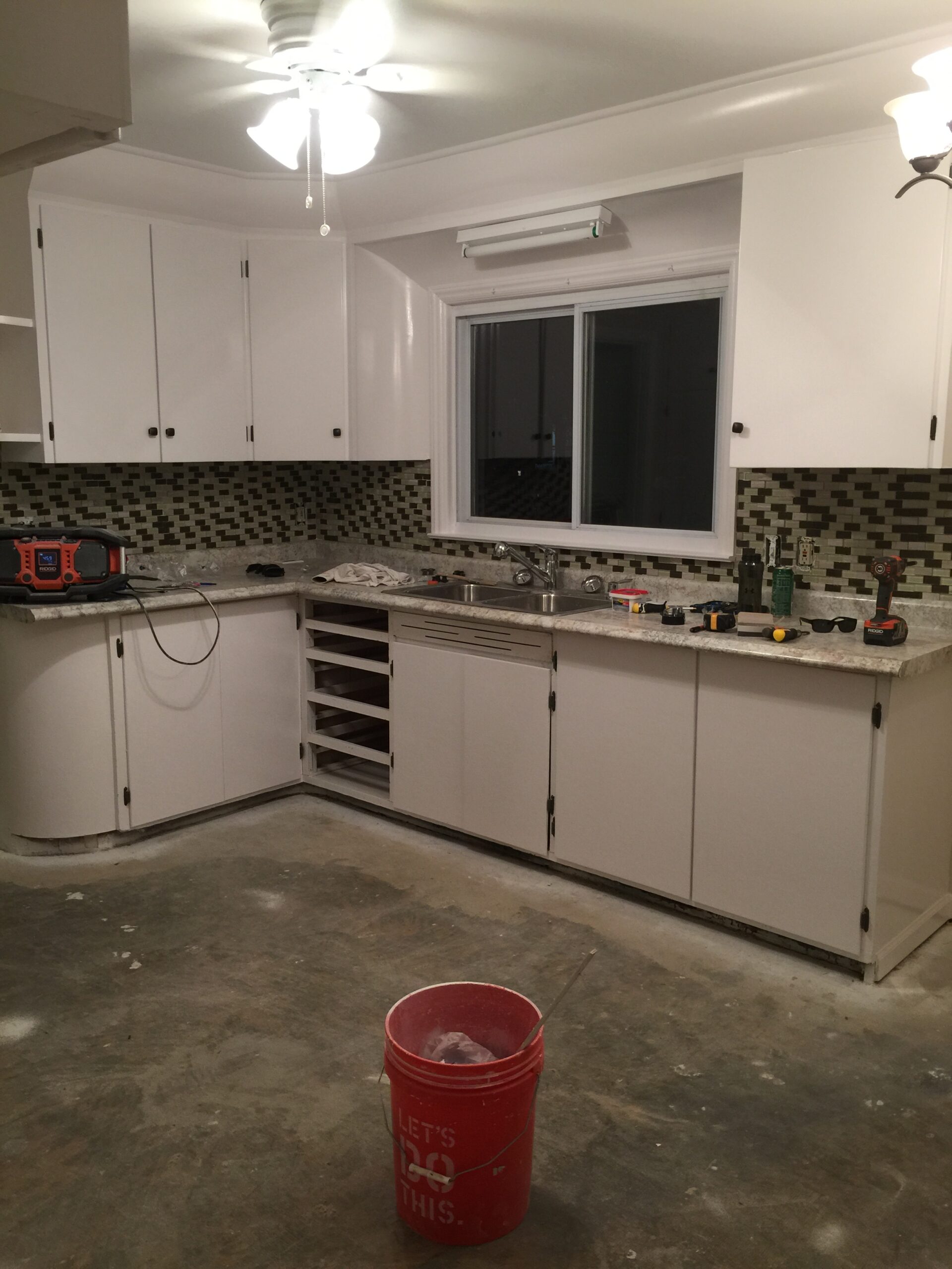 Kitchen with white cabinets, a tile backsplash and a red bucket on the concrete floor during a cabinet refinishing project