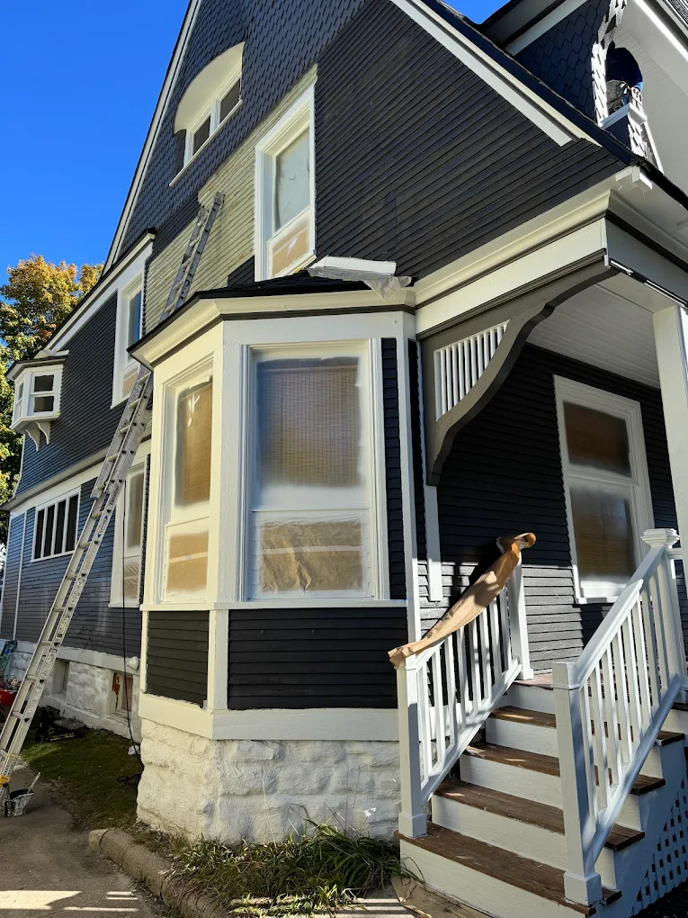Two-story dark blue siding house in Michigan with a bay window under repair, a worker on a ladder and exterior trim being prepared for painting