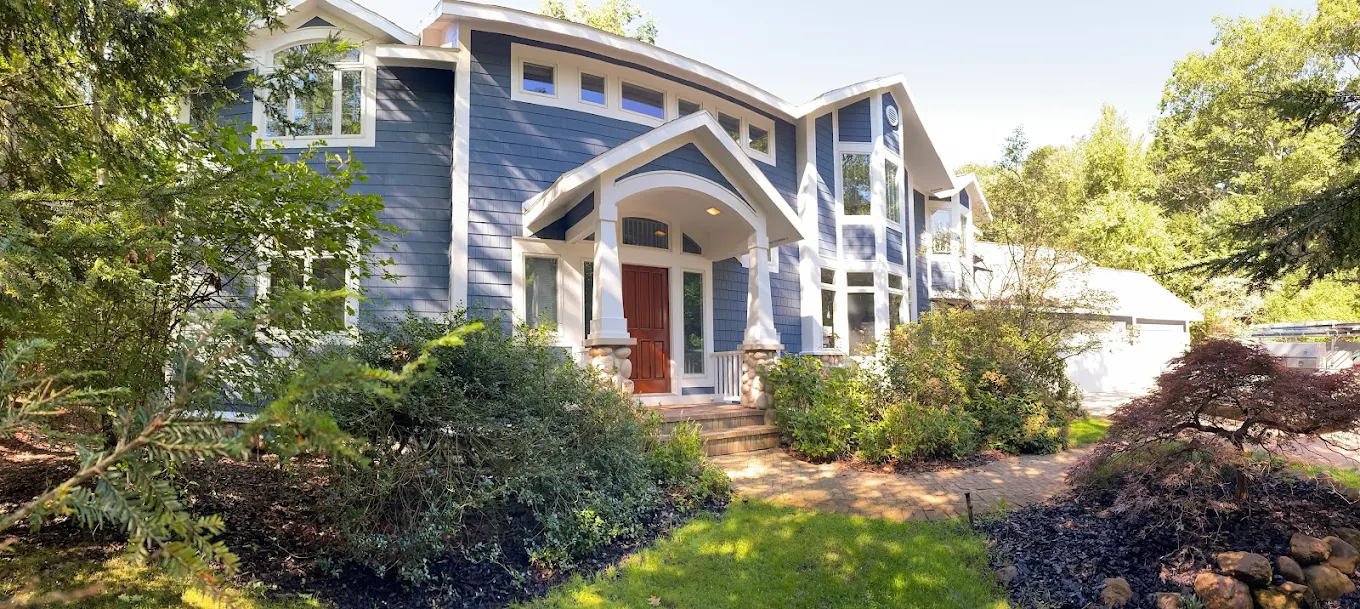 Two-story home with blue siding and dark roof surrounded by trees, shown as an exterior painting prep example